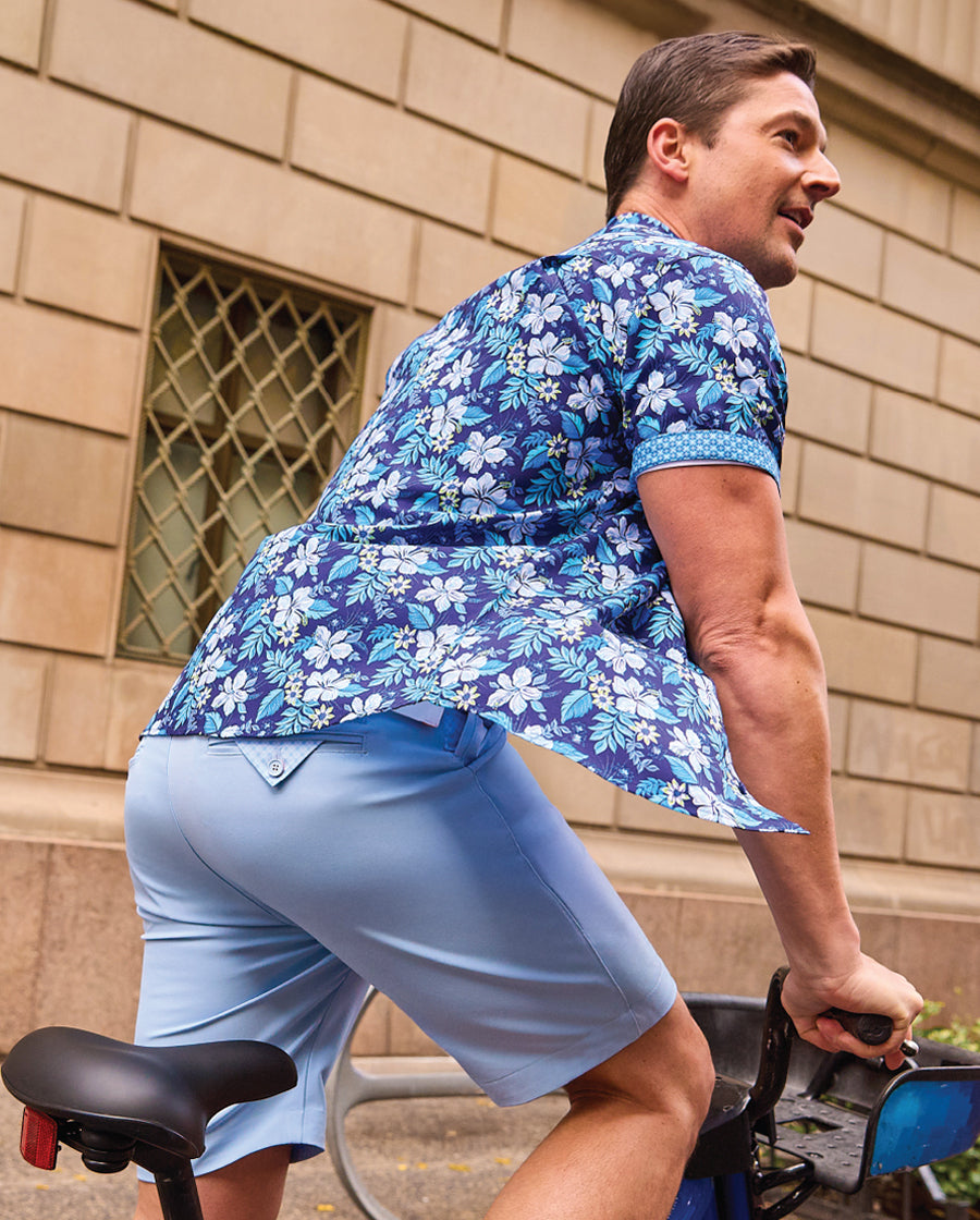 Man wearing a blue floral shirt and light blue shorts riding a bike outdoors.