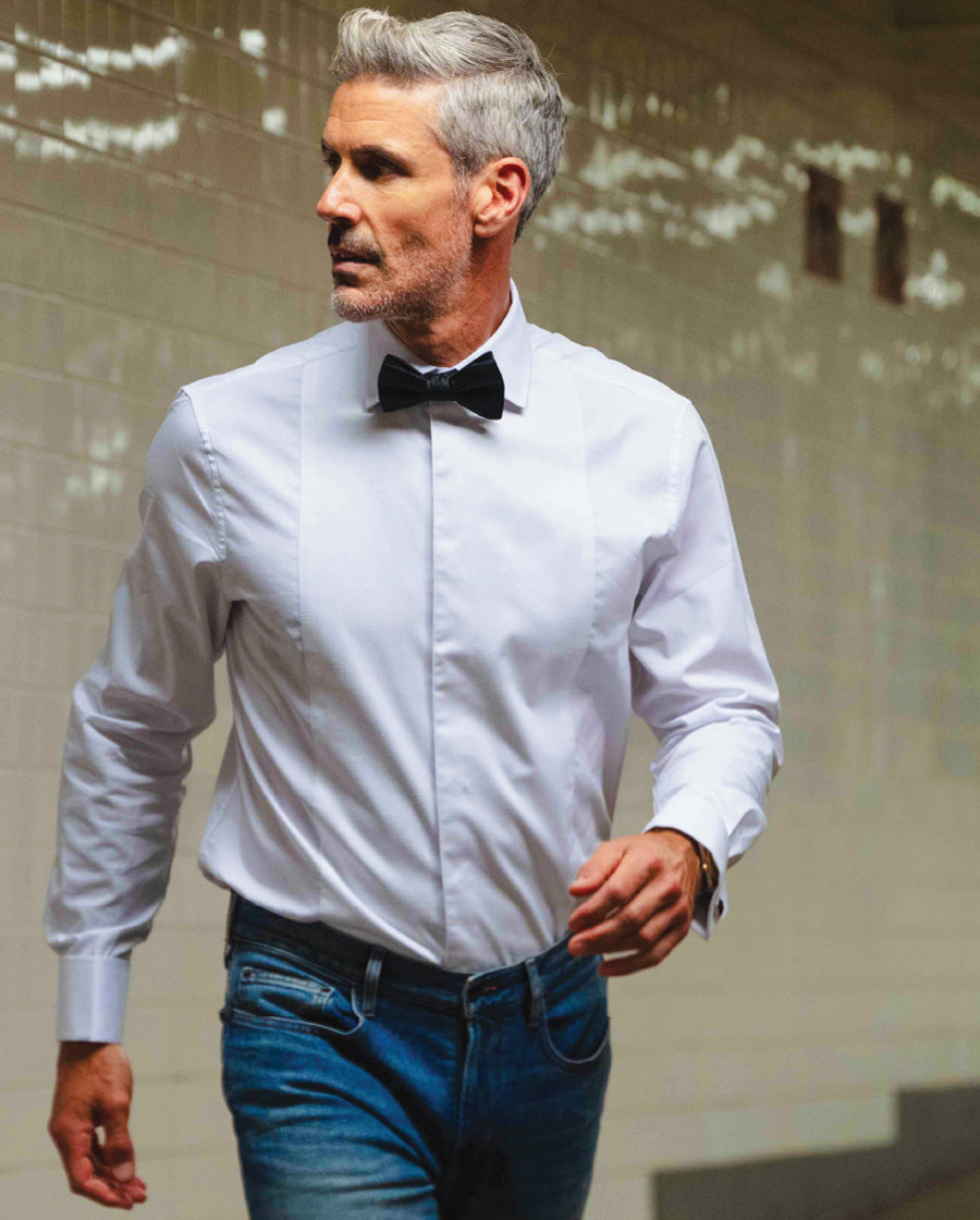 Man wearing a white tuxedo shirt, black bow tie, and blue jeans against a neutral background