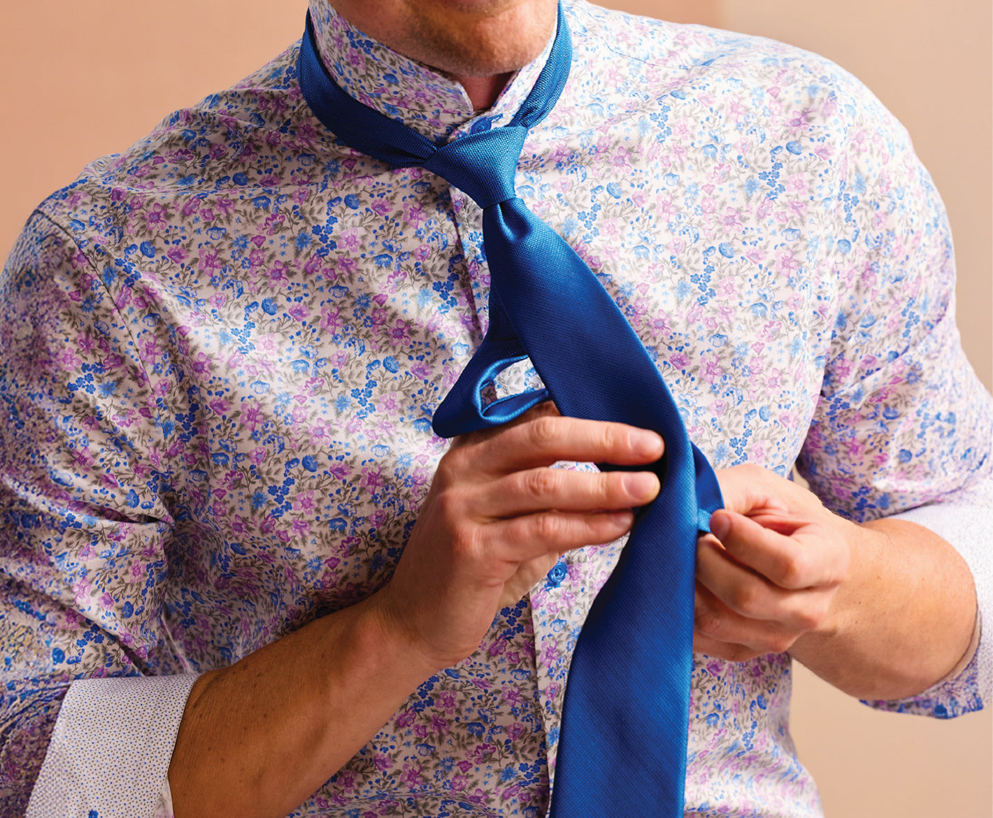 Person adjusting a blue tie against a floral shirt on a beige background