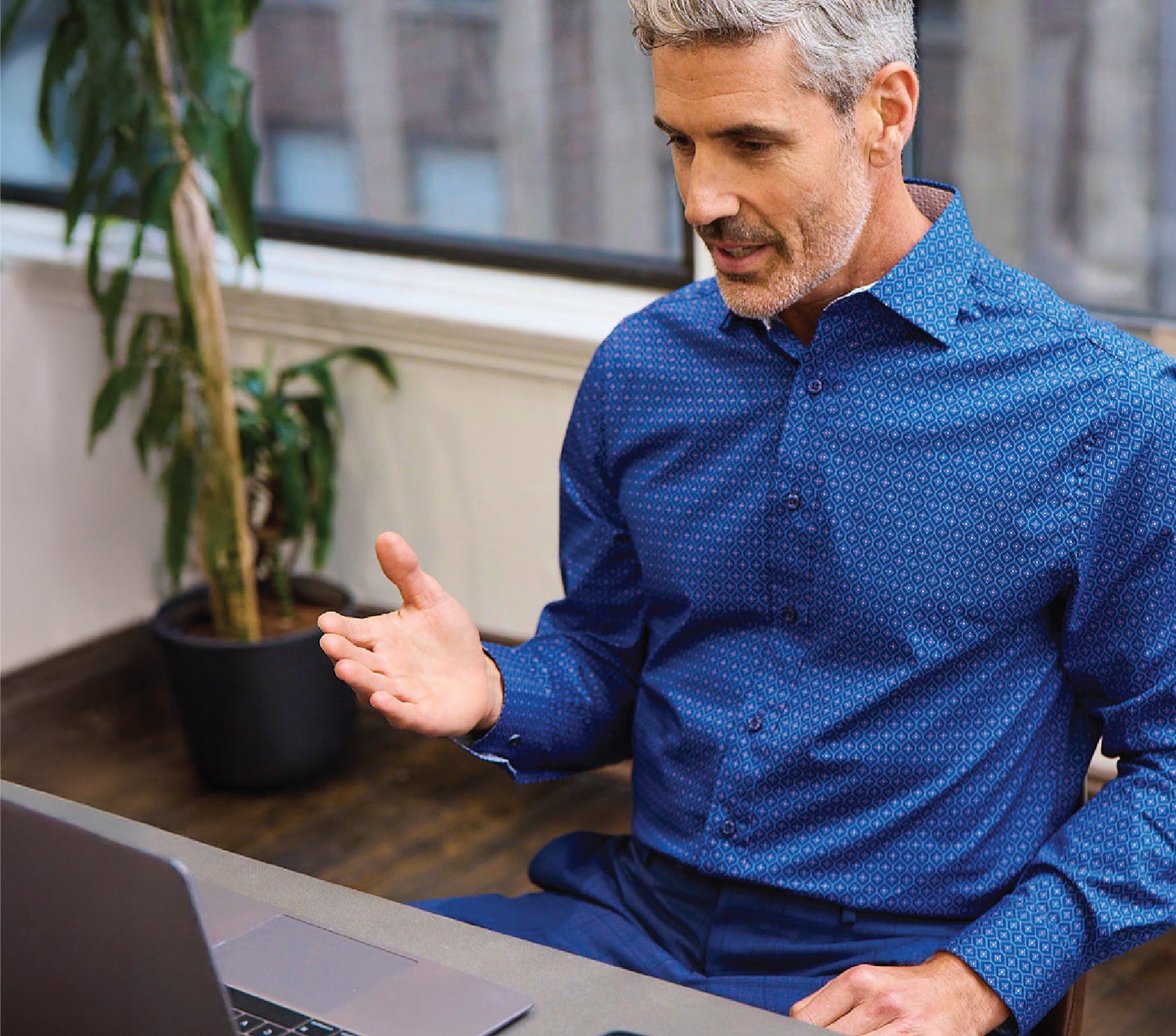 Man in a blue shirt sitting at a desk with a laptop, gesturing as if explaining something.